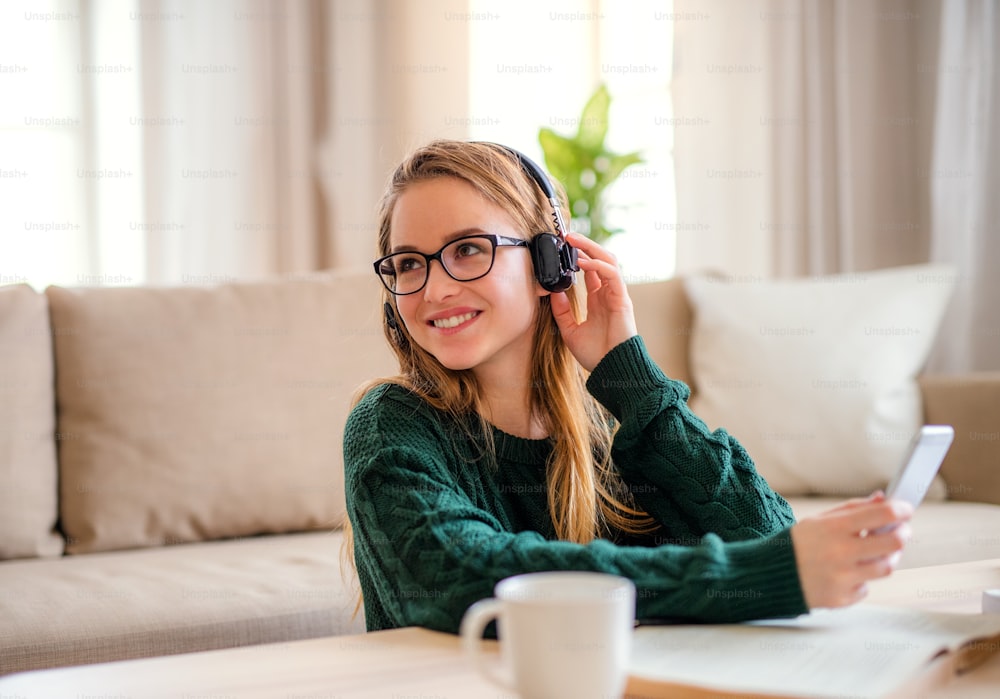 Eine junge, glückliche Studentin, die zu Hause am Tisch sitzt und beim Lernen Kopfhörer benutzt.