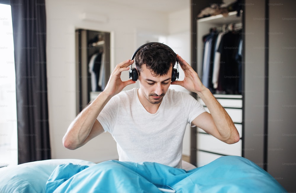 Young man with headphones in bed indoors at home, listening to music.
