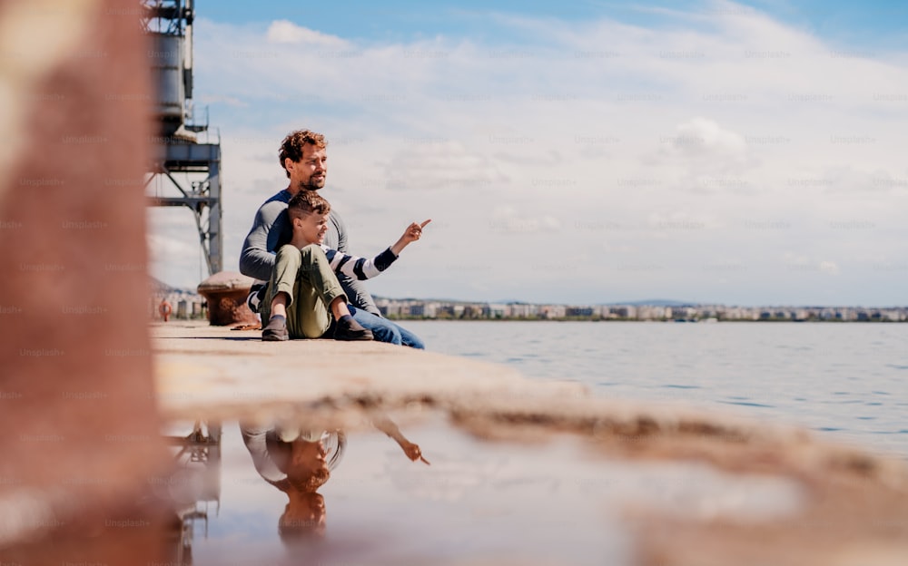 Un padre con un figlio piccolo seduto all'aperto in città in riva al mare, parlando.
