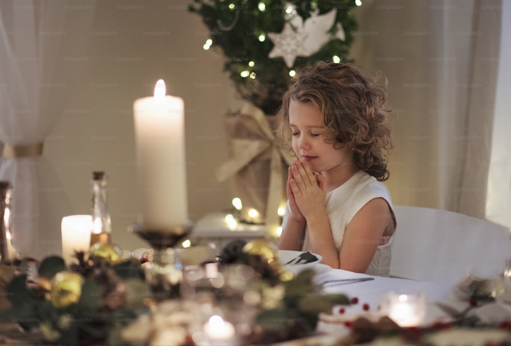 Bambina con gli occhi chiusi e le mani giunte seduta al tavolo al chiuso a Natale, pregando.