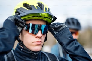 Two mountain bikers standing on road outdoors in winter, putting on sunglasses.