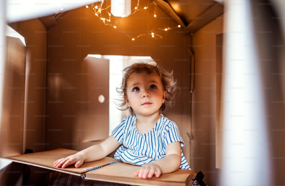 A toddler girl playing indoors in cardboard house at home, looking at photo album.