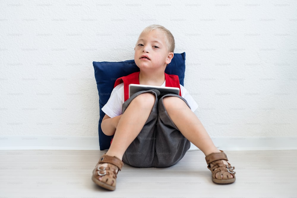 Un ritratto di uno scolaro felice con sindrome di down seduto sul pavimento, usando un tablet.