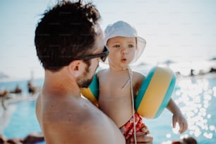 Ein Vater mit kleinem Kind mit Armbinden steht im Sommerurlaub am Schwimmbad.