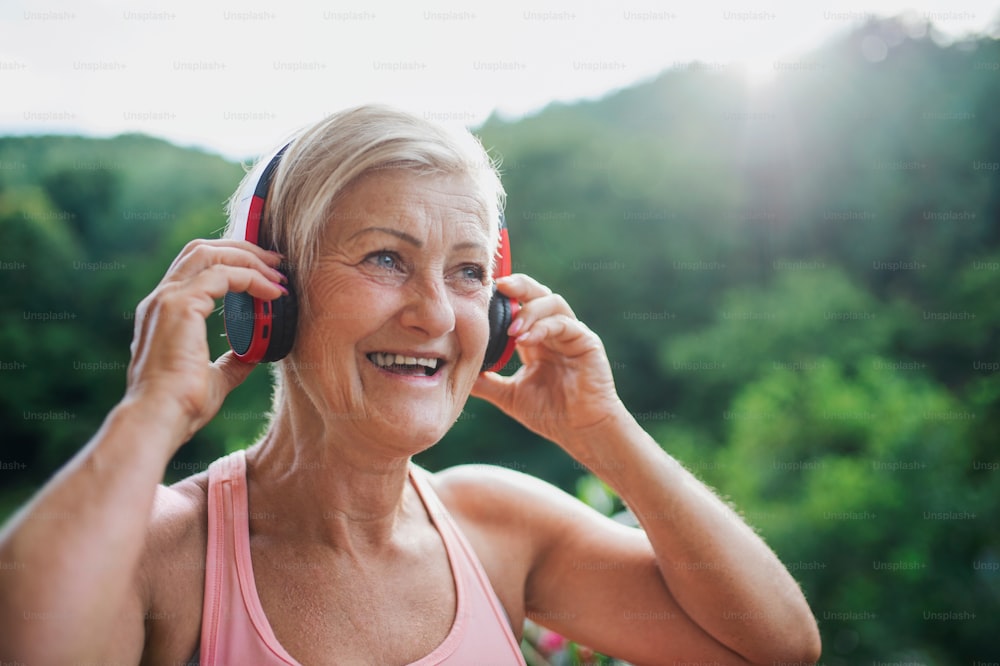 A senior woman with headphones standing outdoors on a terrace in summer, listening to music.