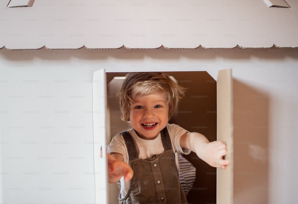 Un petit garçon jouant à l’intérieur avec une maison en carton à la maison, ouvrant la fenêtre.