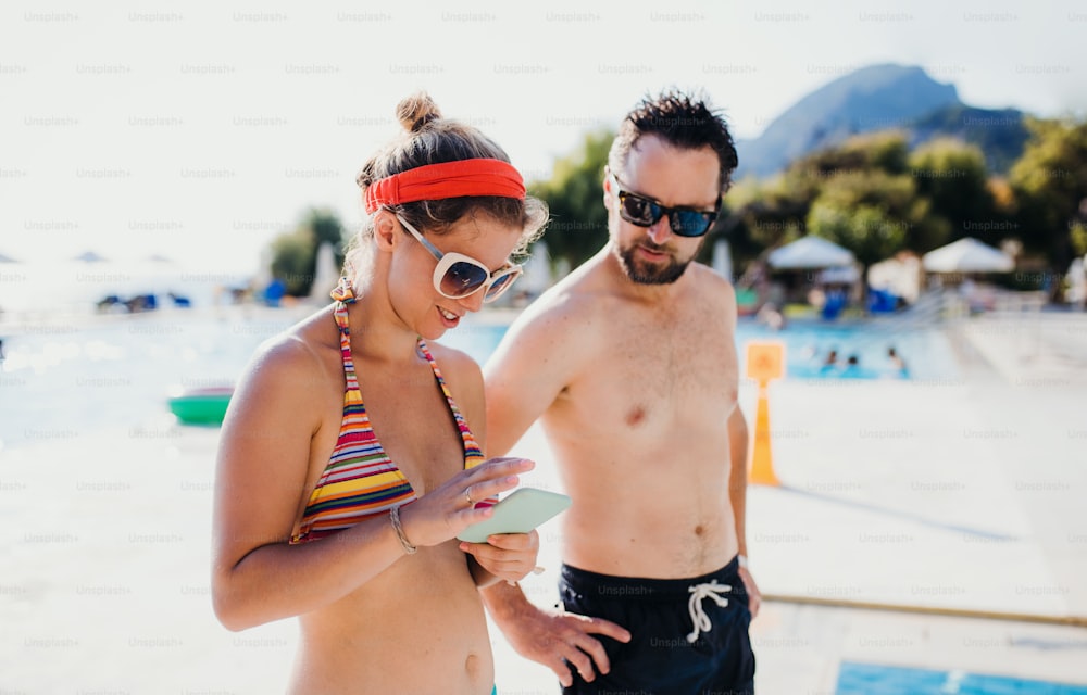 A couple in swimsuit standing by the swimming pool on summer holiday, using smartphone.