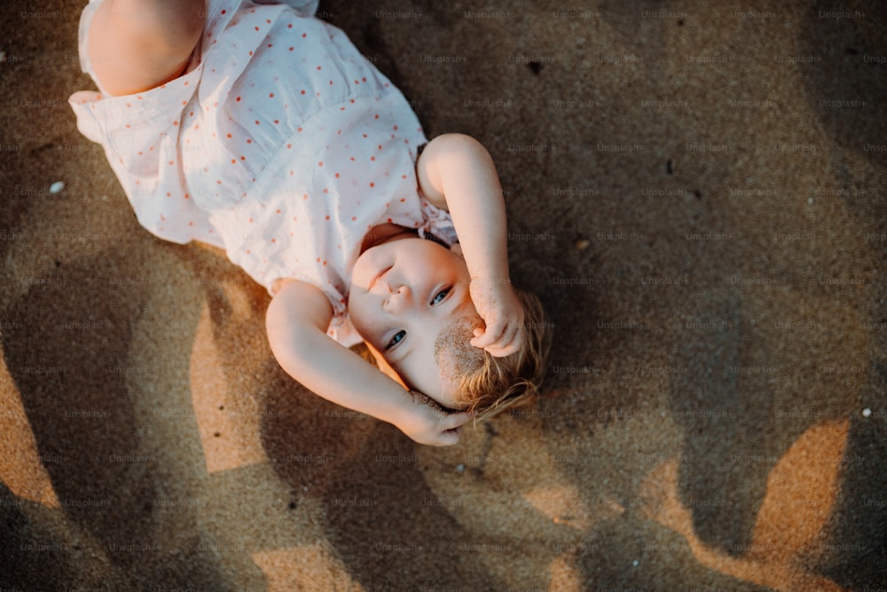Eine Draufsicht auf ein kleines Kleinkind, das im Sommerurlaub am Strand im Sand spielt und in die Kamera schaut.