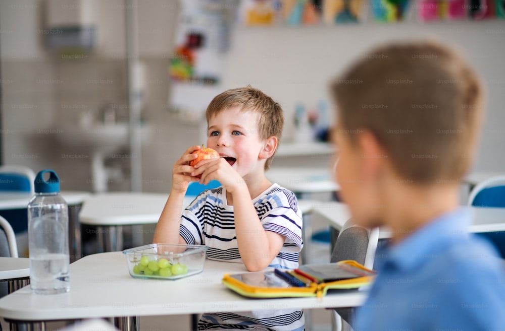 Kleine Schulkinder sitzen am Schreibtisch im Klassenzimmer und essen Obst als Snack.
