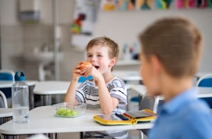 Kleine Schulkinder sitzen am Schreibtisch im Klassenzimmer und essen Obst als Snack.