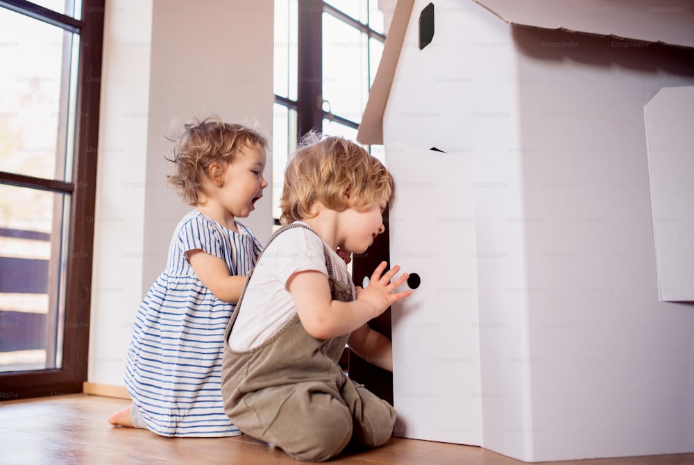 Two toddler children playing with a carton paper house indoors at home. Copy space.