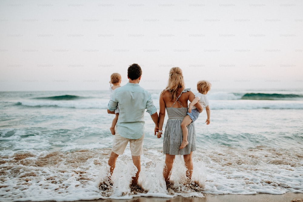 Uma visão traseira da família jovem com duas crianças pequenas em pé na praia nas férias de verão.