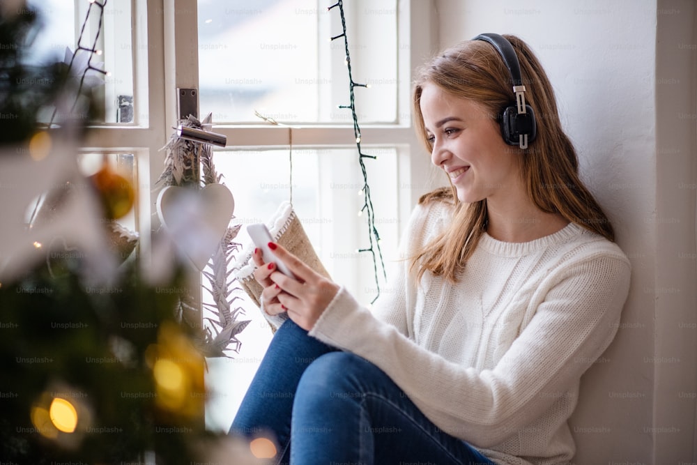 Portrait of young woman sitting indoors at home at Christmas, using smartphone and headphones.