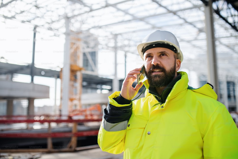 Ingeniero hombre maduro de pie en el sitio de construcción, usando un teléfono inteligente.