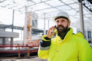 Mature man engineer standing on construction site, using smartphone.