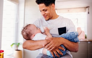 A portrait of cheerful father bottle feeding a small toddler son indoors at home.