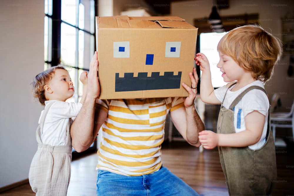 Dos niños pequeños felices con un padre y un monstruo de cartón jugando en el interior de casa.