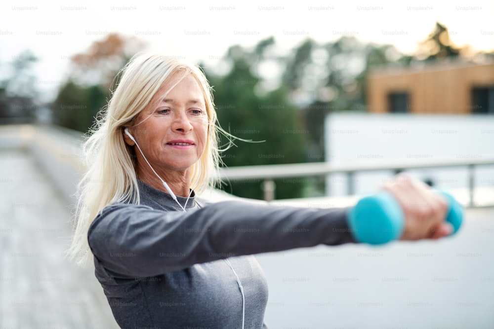 A senior woman with earphones and dumbbells outdoors doing exercise on terrace.