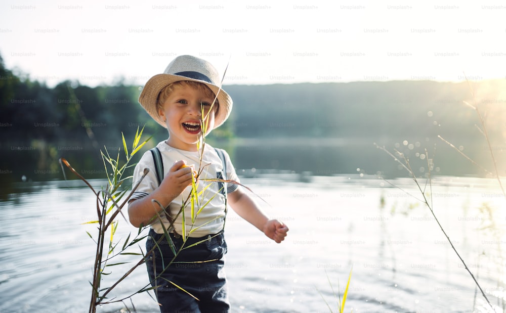 Un piccolo bambino con un cappello in piedi vicino a un lago al tramonto. Copia spazio.