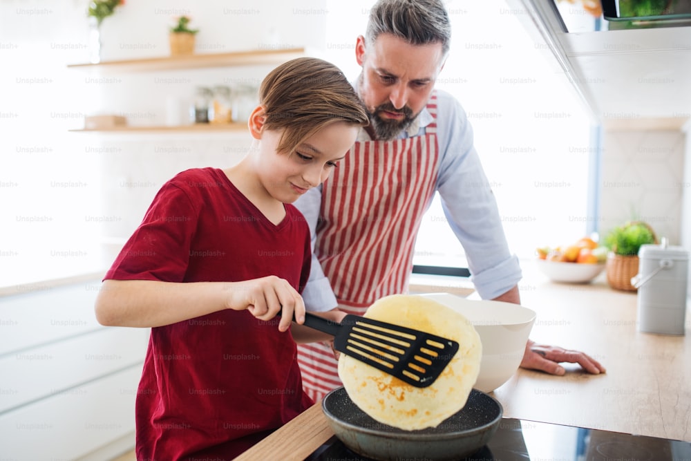 Ein reifer Vater mit kleinem Sohn drinnen in der Küche, der Pfannkuchen macht.