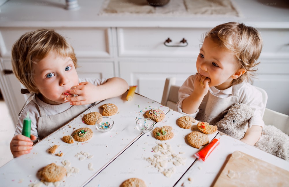 Two happy small toddler children sitting at the table, decorating and eating cakes at home.