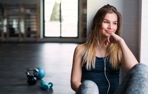 A portrait of young girl or woman earphones sitting in a gym, listening to music. Copy space.