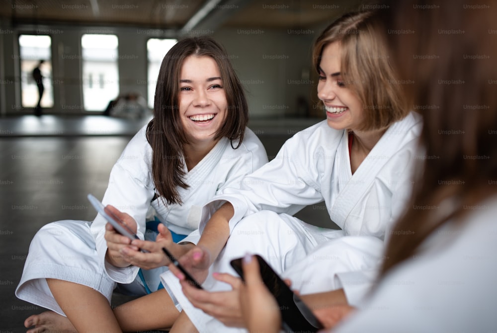 Un groupe de jeunes femmes karatés avec des smartphones à l’intérieur d’un gymnase, se reposant.