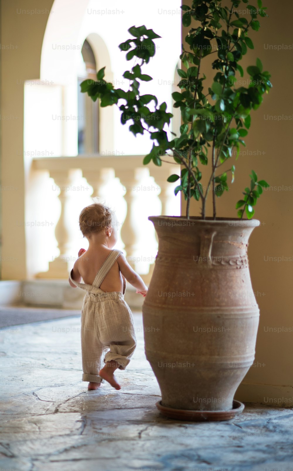 A rear view of small toddler girl walking around a big flower pot on a terrace on summer holiday.