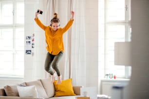 Uma jovem estudante com fones de ouvido e telefone fazendo uma pausa ao estudar, pulando no sofá.