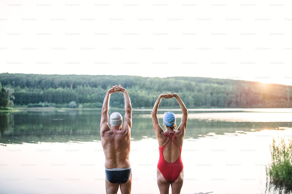 Vista trasera de una pareja mayor en traje de baño estirándose junto al lago al aire libre antes de nadar,
