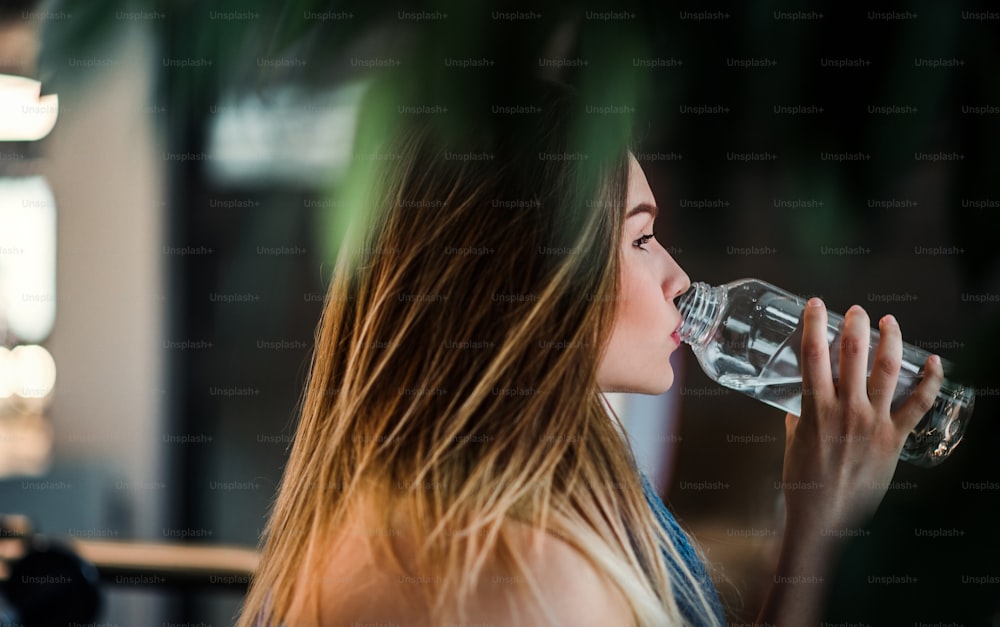 어린 소녀 또는 여자가 체육관에 서서 병에서 물을 마시는 측면보기.