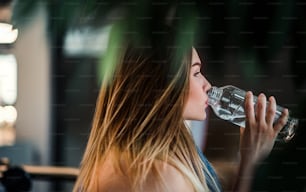 Una vista laterale di una giovane ragazza o donna in piedi in una palestra, bevendo acqua da una bottiglia.