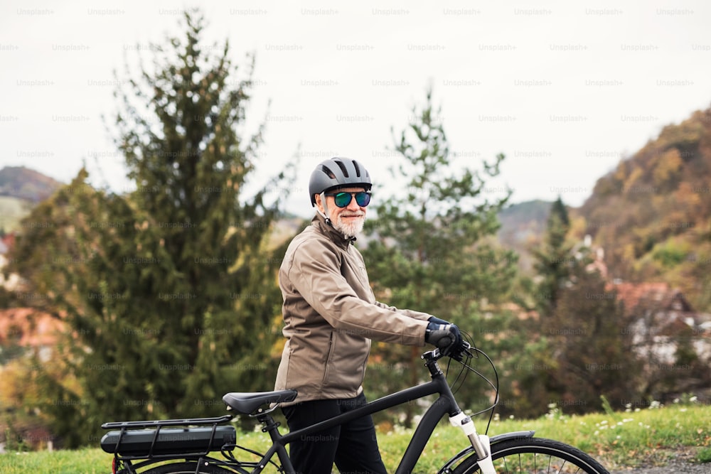 Un homme âgé actif avec un casque, des lunettes de soleil et un vélo électrique debout à l’extérieur sur une route dans la nature.