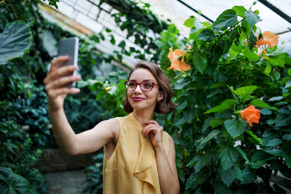 Vista frontal de una mujer joven con un teléfono inteligente de pie en el jardín botánico, tomando una selfie.