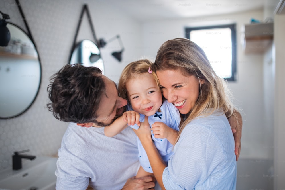 Uma jovem família com filha pequena dentro de casa no banheiro, abraçando.