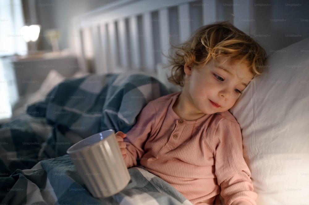 Small sick toddler girl lying in bed indoors at home, holding cup with tea.