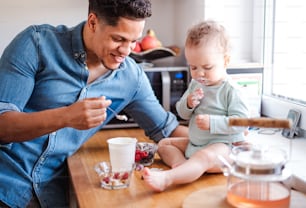 Um pai e um filho pequeno comendo frutas e iogurte na cozinha dentro de casa.
