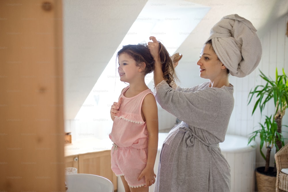 Porträt der schwangeren Frau mit kleiner Tochter drinnen im Badezimmer zu Hause, kämmt Haare.