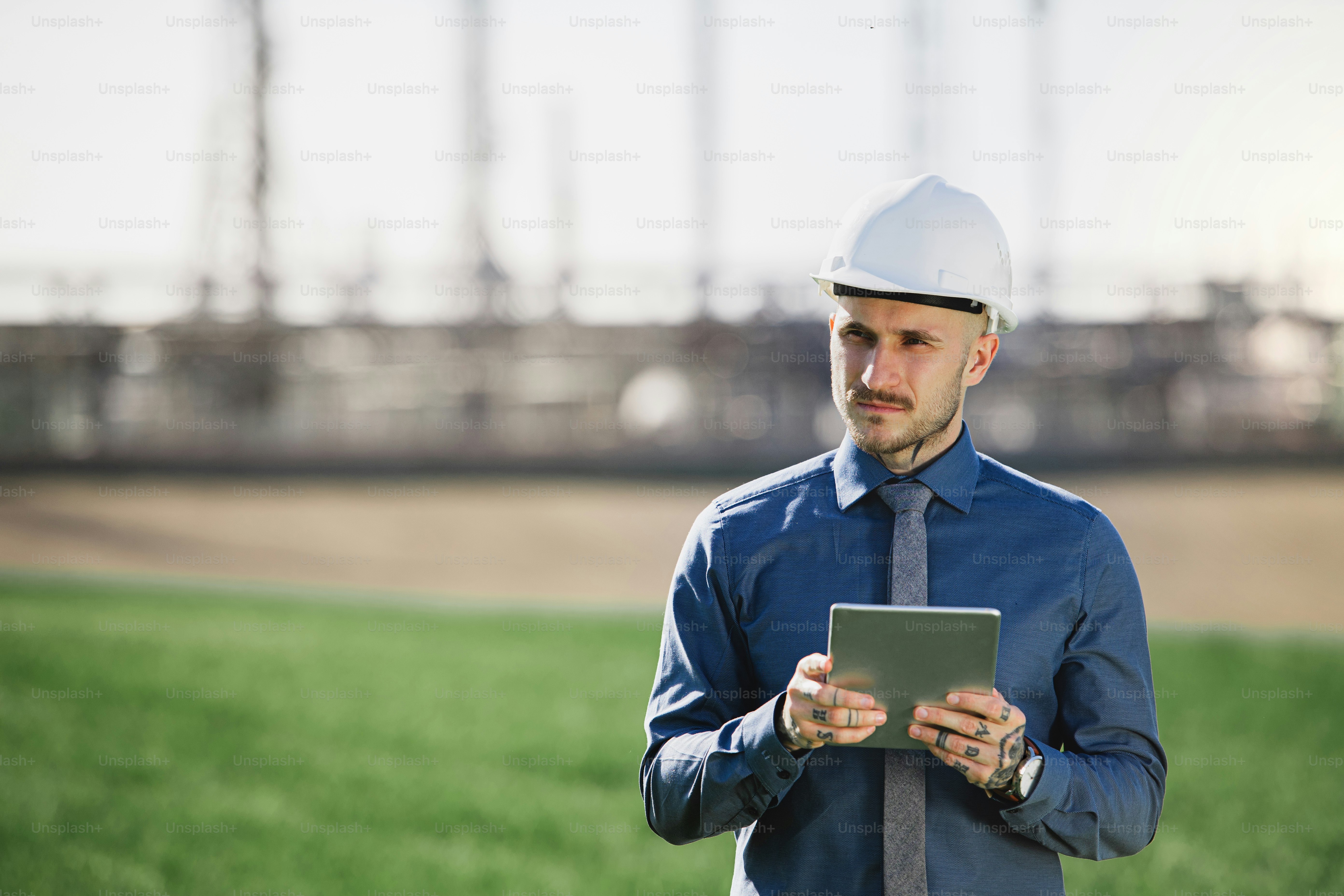 Young engineer with hard hat and tablet standing outdoors by oil refinery. Copy space.