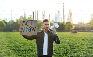 Portrait of young activist with placard and megaphone standing outdoors by oil refinery, protesting.