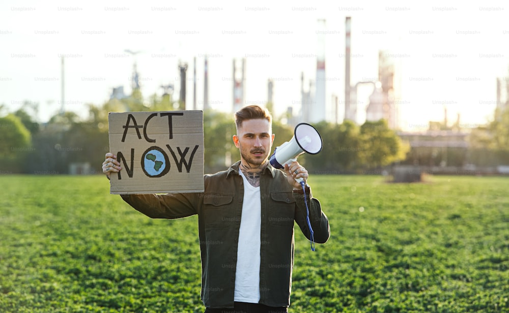 Retrato de un joven activista con pancarta y megáfono de pie al aire libre junto a una refinería de petróleo, protestando.