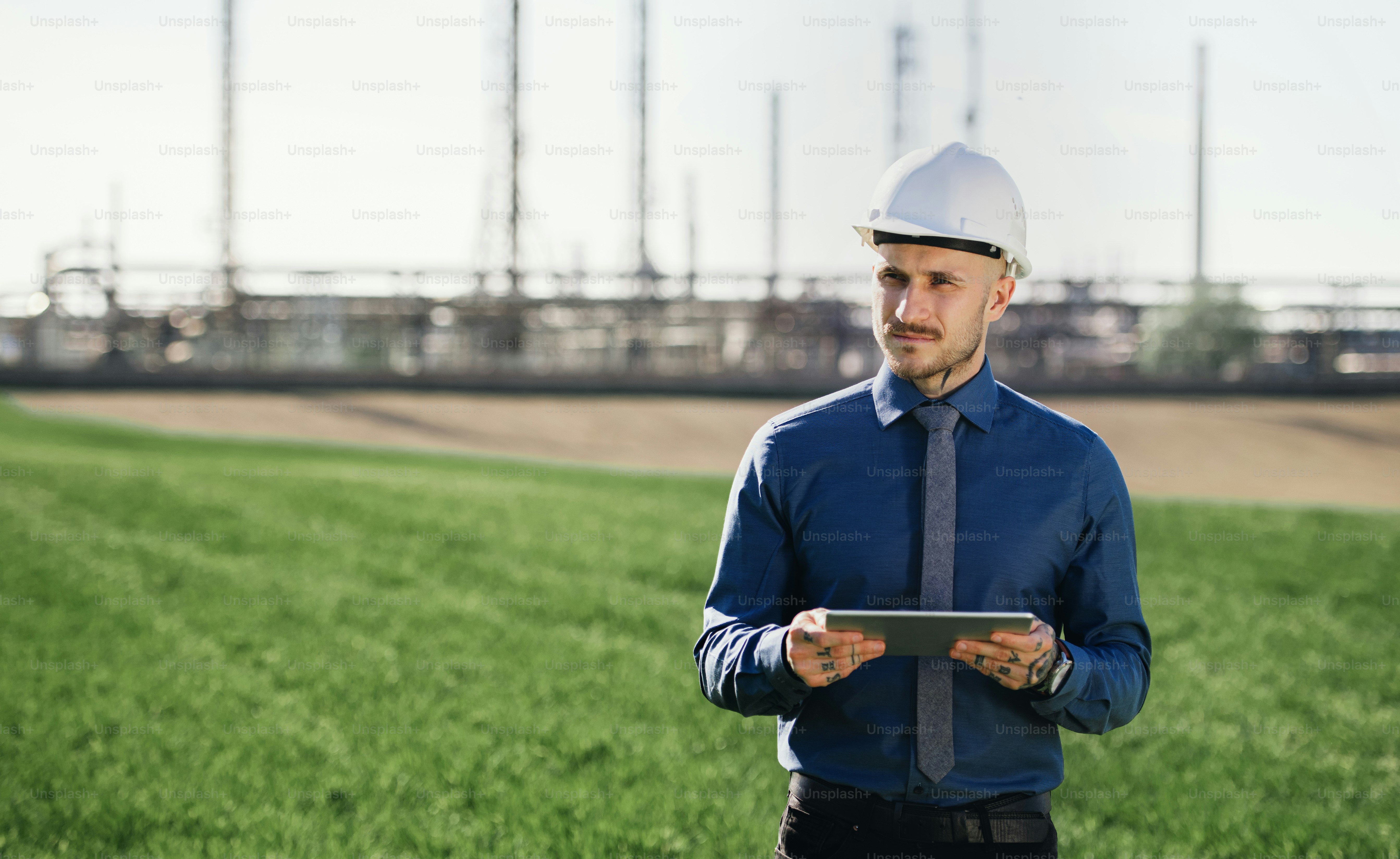 Young engineer with hard hat and tablet standing outdoors by oil refinery. Copy space.