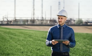 Young engineer with hard hat and tablet standing outdoors by oil refinery. Copy space.