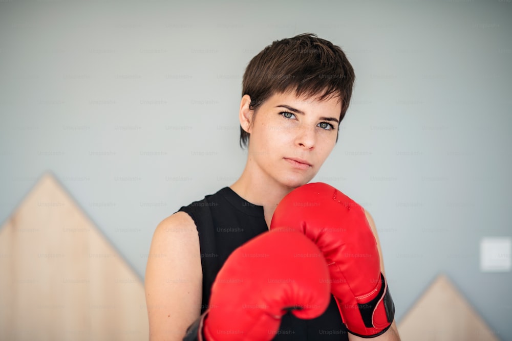 Front view of young woman with red boxing gloves standing indoors at home.