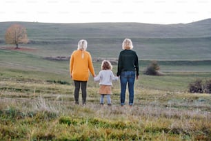 Rear view of small girl with mother and grandmother on a walk in autumn nature, holding hands.