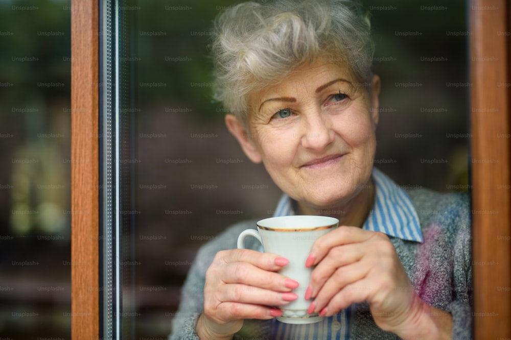 Mujer mayor triste con café mirando hacia el interior de su casa, coronavirus y concepto de cuarentena.