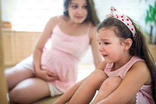 Portrait of pregnant woman with crying small daughter indoors at home, sitting.