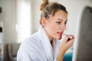 A young woman indoors at home in the morning, putting on lipstick.