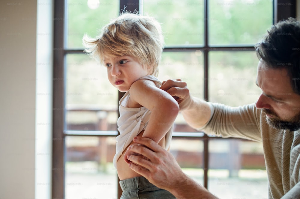 Father with small sick unhappy son indoors at home, checking his hurt back.