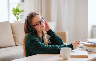 A young happy college female student sitting at the table at home, using headphones when studying.